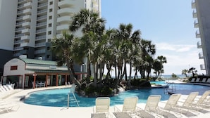 A resort pool area with lounge chairs, palm trees, and a building with multiple balconies. - 3BD/3BA Tower 3 Corner Renovated Condo! Comes with beach chairs and umbrella! (Panama City Beach)