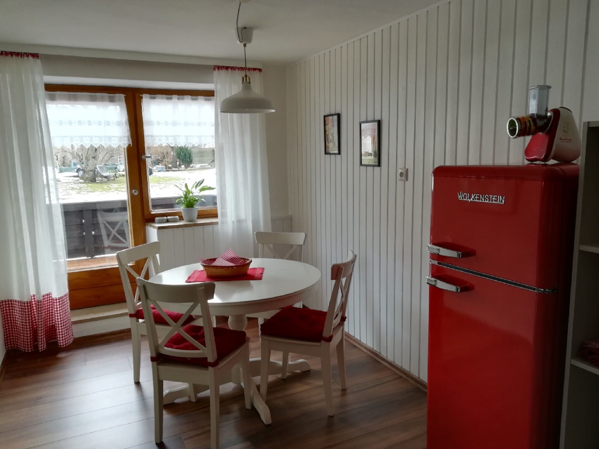 A dining area with a round table and four chairs, a red refrigerator, and a window with curtains.