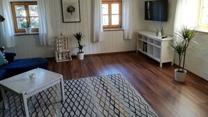 Un soggiorno con pavimento in legno, un divano blu, un tavolino da caffè bianco, un tappeto decorato, una televisione e due finestre con tende.