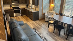 Una cocina moderna con armarios de madera oscura, electrodomésticos de acero inoxidable y un horno empotrado. Hay un comedor con una mesa redonda y sillas, y una zona de estar acogedora con un sofá de cuero.