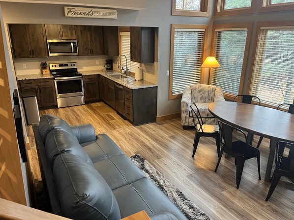 A modern kitchen with dark wood cabinets, stainless steel appliances, and a built-in oven. There is a dining area with a round table and chairs, and a cozy seating area with a leather couch.