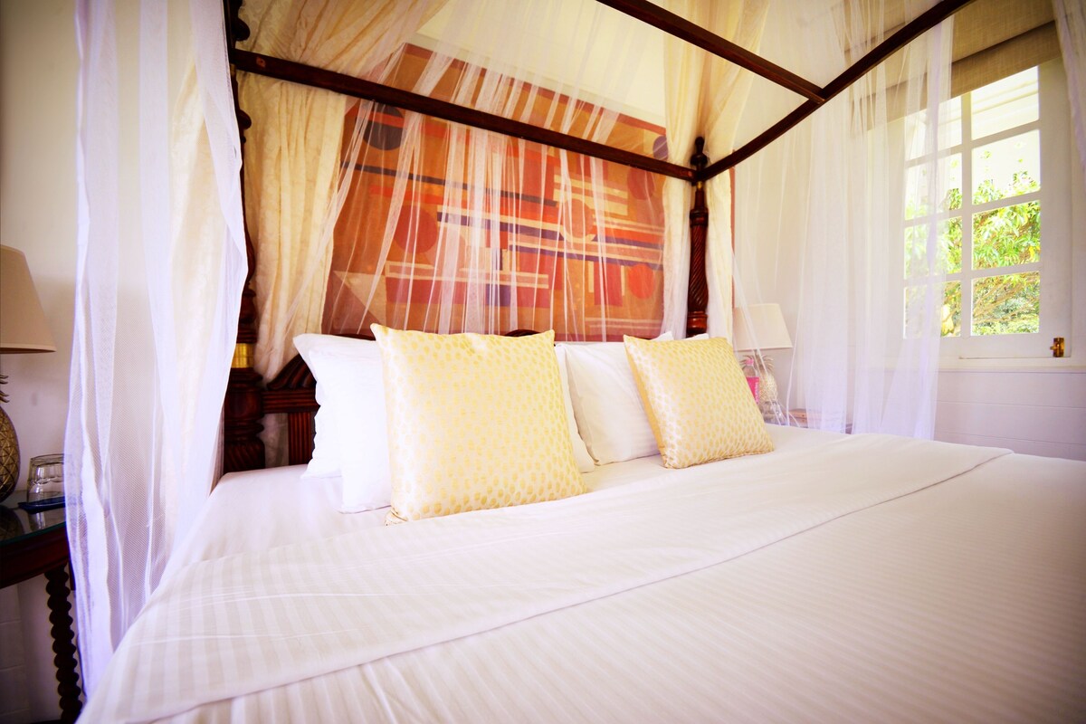 A four-poster bed with a canopy, a bedside table with a lamp, and a window with curtains.