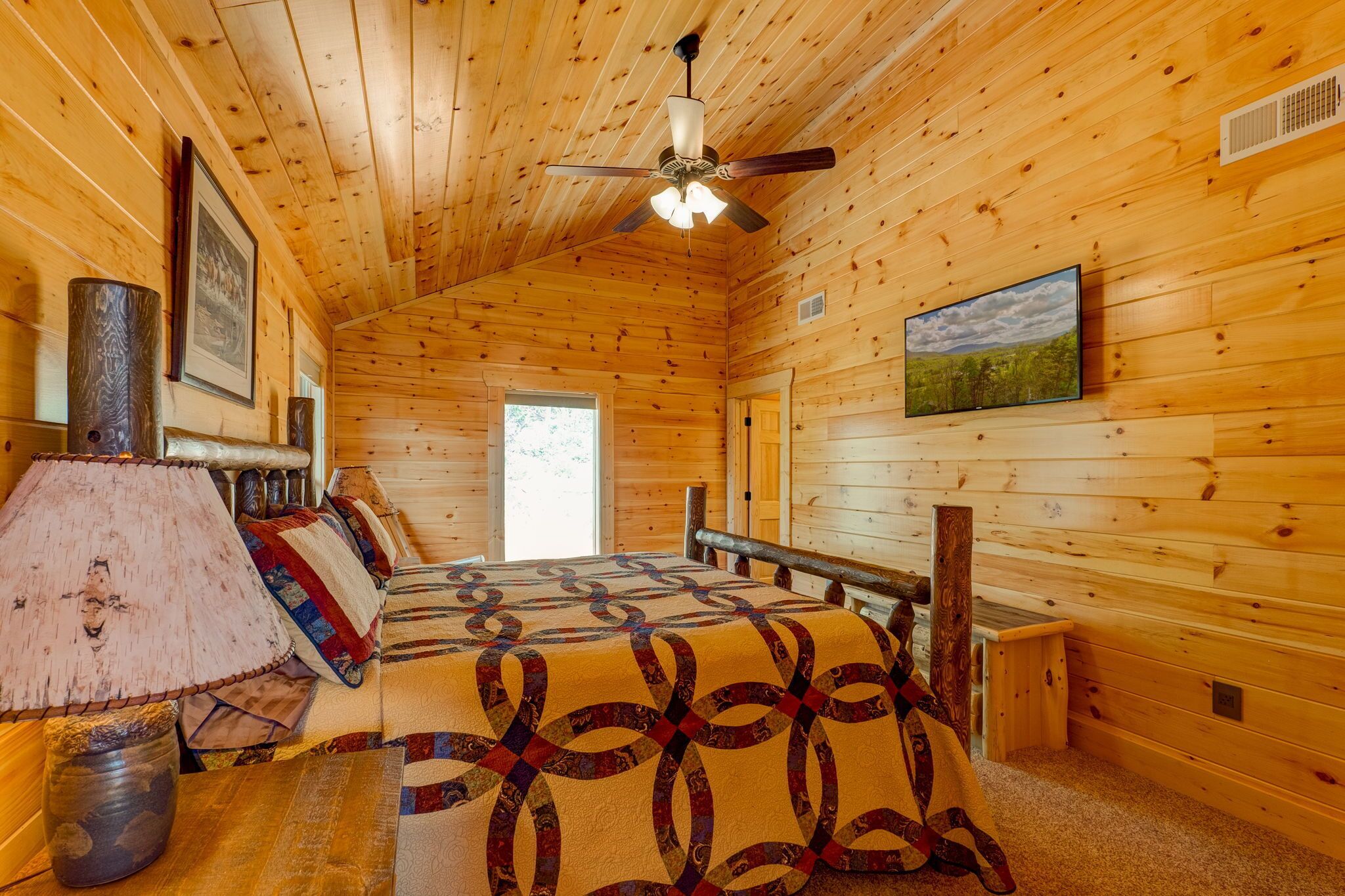 Une chambre rustique en bois, avec un lit, un ventilateur de plafond, une lampe et un tableau encadré au mur.