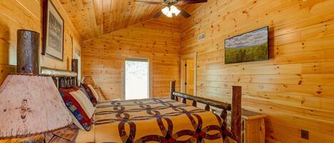 Une chambre rustique en bois, avec un lit, un ventilateur de plafond, une lampe et un tableau encadré au mur.