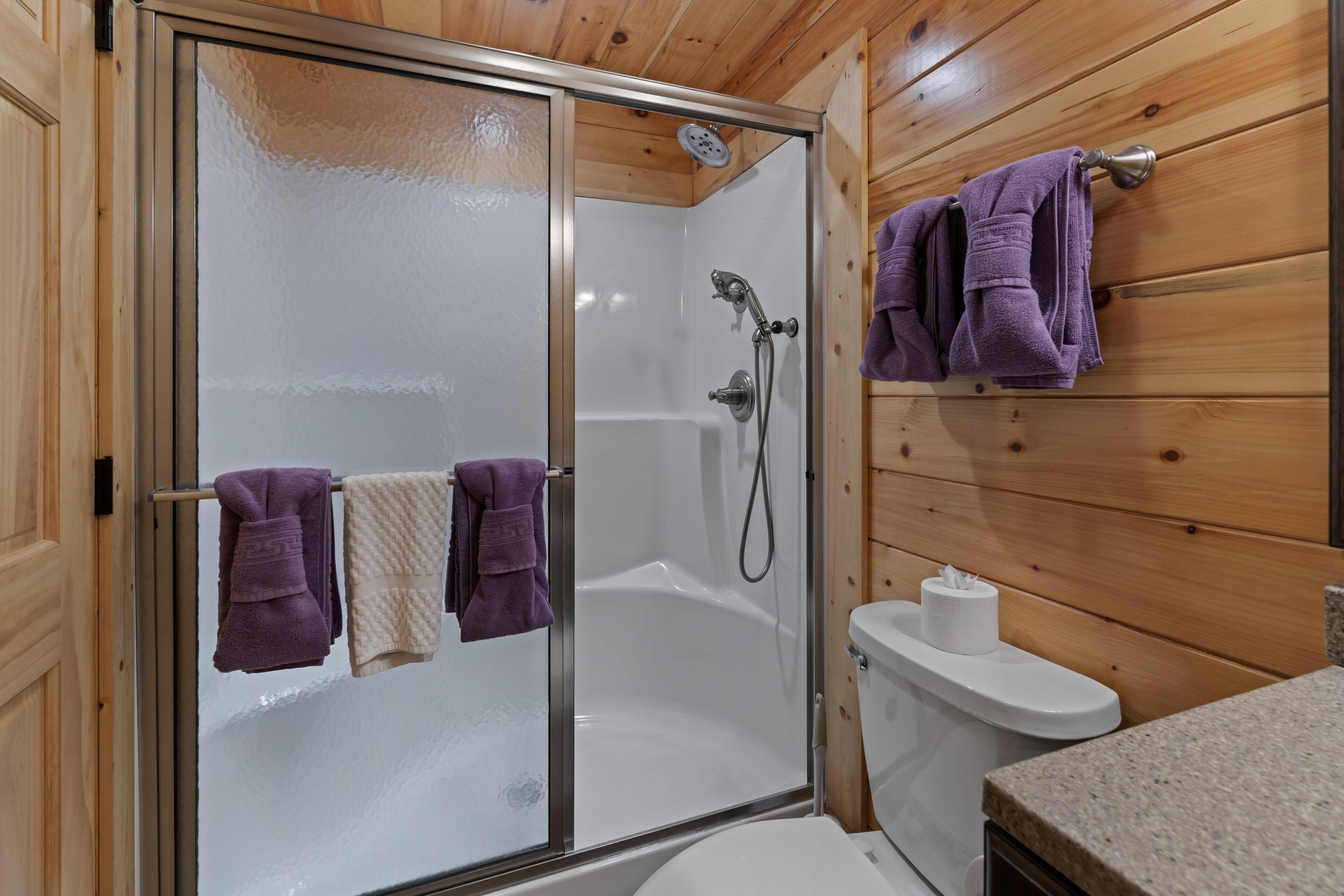 Een badkamer met houten lambrisering, een douchecabine van glas, twee handdoeken en een toilet.