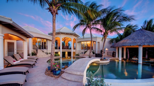 A resort with a swimming pool, lounge chairs, palm trees, and a thatched-roof structure.