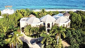 A large, white, multi-story building with a dome roof, surrounded by lush greenery and a beach view.