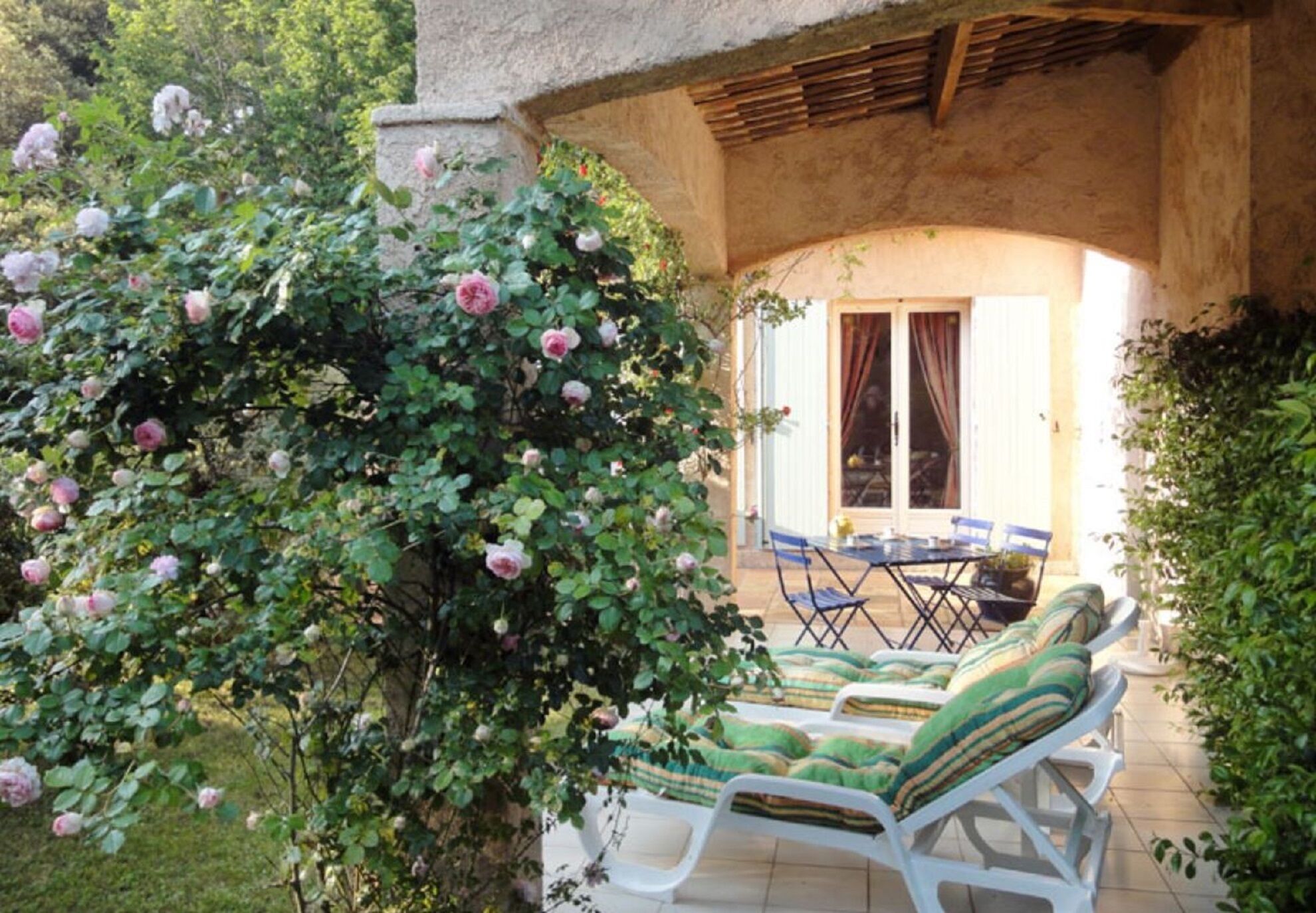 A patio with white lounge chairs and a table, surrounded by lush greenery and flowering bushes.