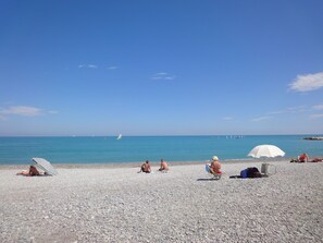 Une plage de galets où des gens se prélassent au soleil, et un voilier à l’horizon.