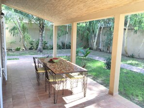 Une terrasse couverte avec un sol carrelé, une table avec des chaises et une pastèque posée sur la table.