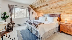 Une chambre avec un plafond en bois, un lit recouvert de draps blancs, une table de chevet, une chaise et une fenêtre avec des rideaux.