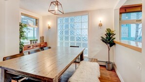 Un coin repas comprenant une table en bois, des chaises, un luminaire suspendu, une grande fenêtre avec du verre dépoli et une plante en pot.