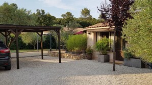 Un espace extérieur abrité, doté d’un sol en gravier, une voiture garée sous une pergola et un bâtiment avec un toit en tuiles et des plantes en pot.