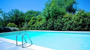 A clear blue swimming pool with metal ladders on the left, surrounded by lush green trees and a clear sky.