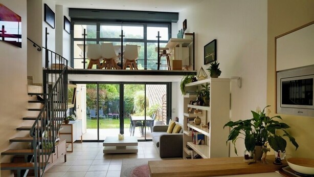A modern living room with a staircase, a dining area with chairs, a sofa, and a kitchen area with a counter and plants.