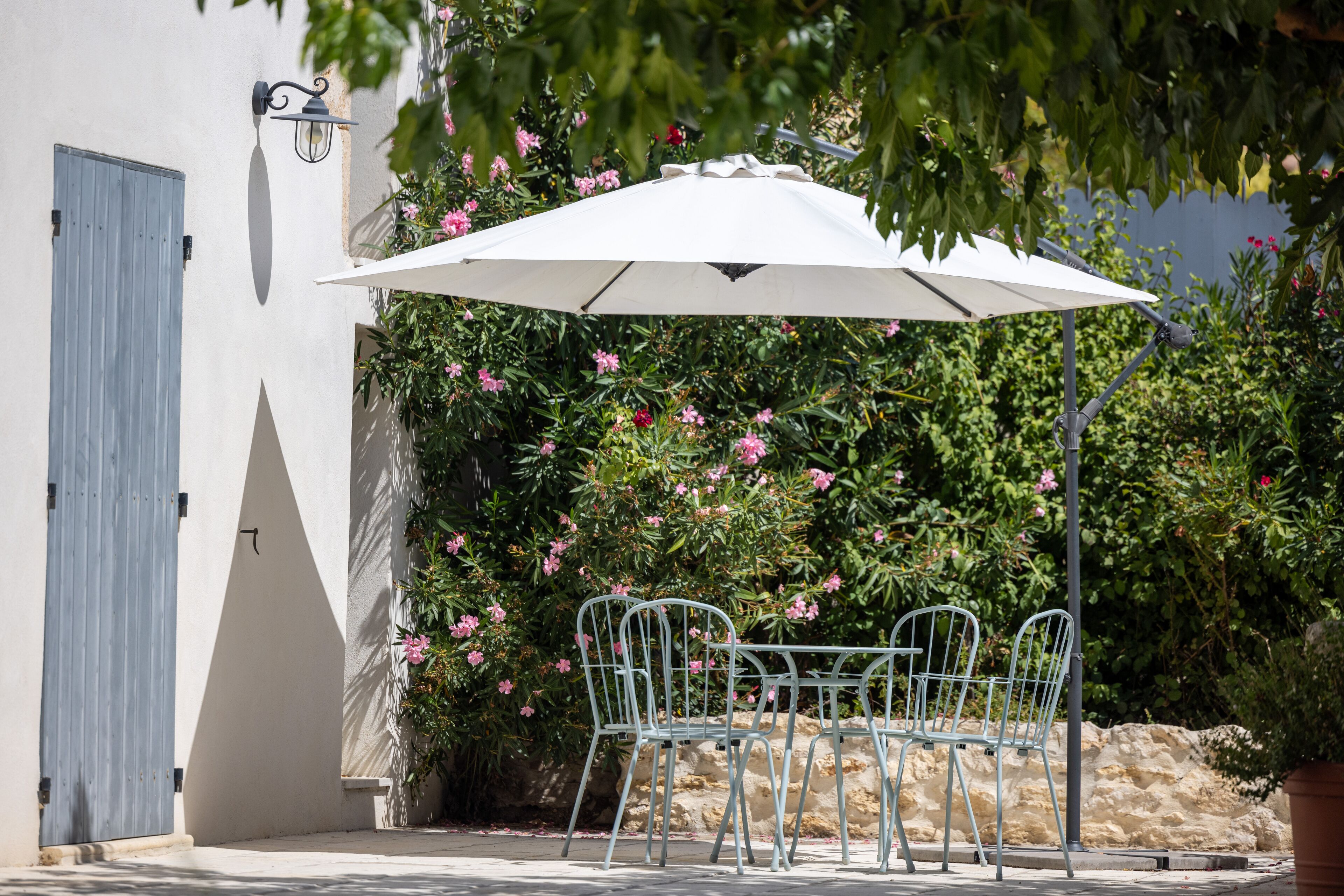Une terrasse avec une table et des chaises sous un parasol blanc, entourée de verdure et d’une porte bleue.