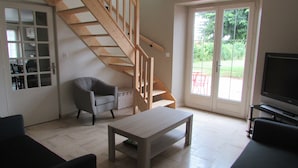 A living room with a wooden staircase, a grey armchair, a wooden coffee table, a TV, and a sliding glass door leading to an outdoor area.