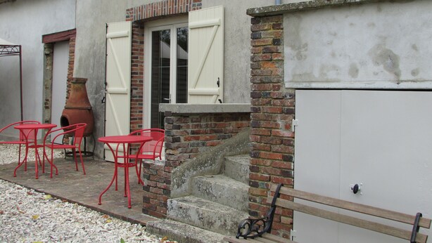 A building with a brick facade, a set of outdoor steps leading to a door with white shutters, and red metal chairs and tables arranged outside.
