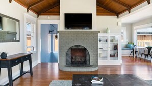 A spacious living room with a fireplace, wooden flooring, a television mounted above the fireplace, and a white cabinet with glass doors.