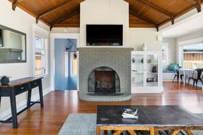 A spacious living room with a fireplace, wooden flooring, a television mounted above the fireplace, and a white cabinet with glass doors.