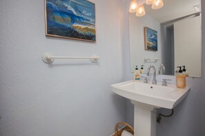 Um banheiro com uma pia branca, duas torneiras e um espelho. Há duas pinturas na parede, um porta-toalhas e uma cesta.
