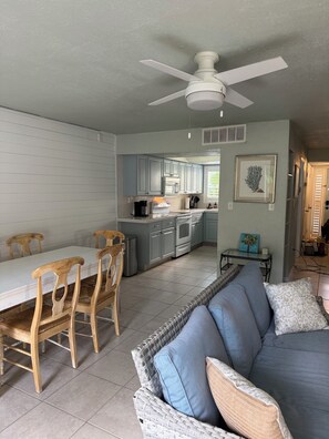 A dining area with a white table and wooden chairs, a kitchen with light blue cabinets, and a ceiling fan.