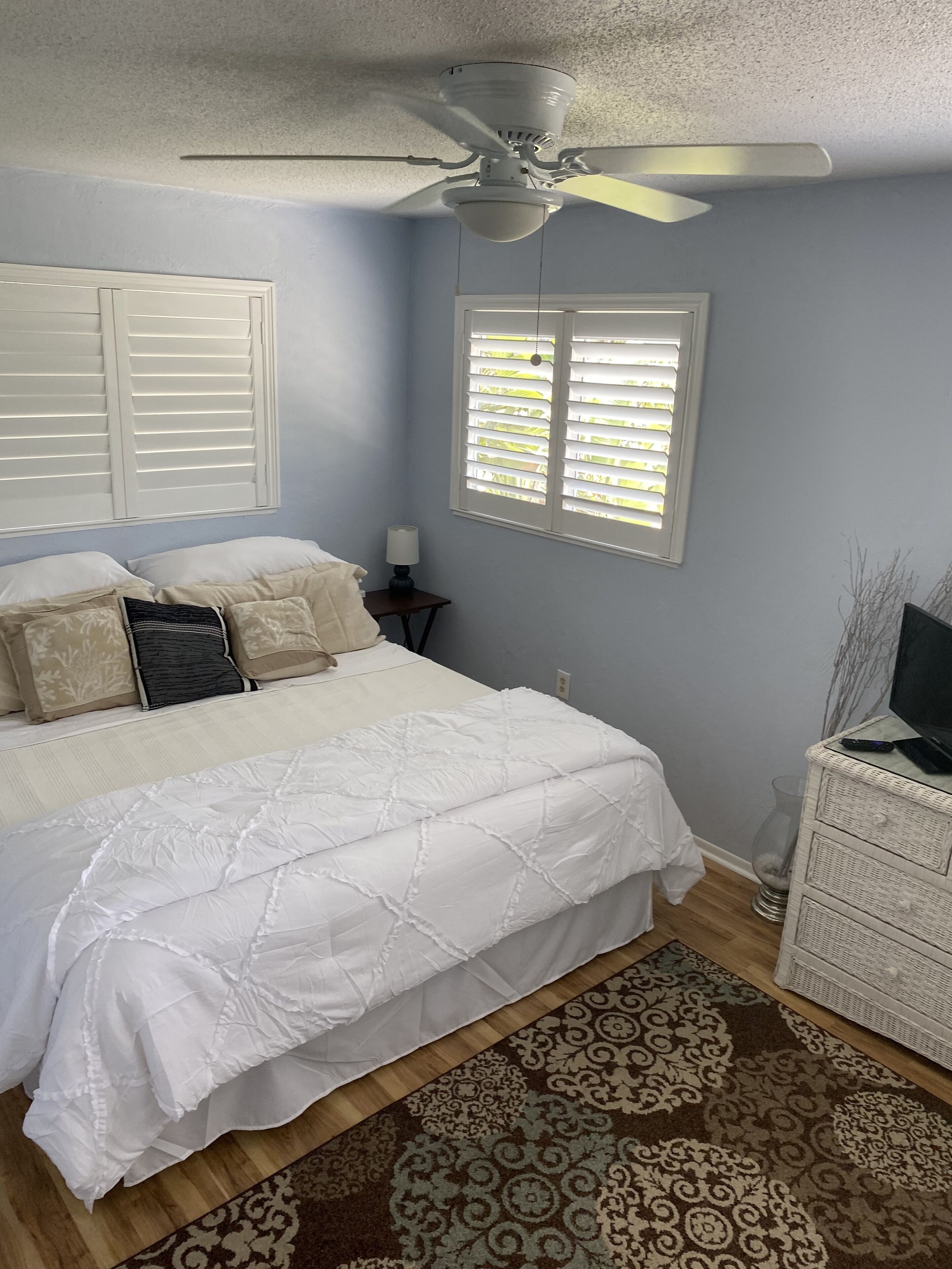 Un dormitorio con cama, ventilador de techo, ventana con persianas, mesita de noche con lámpara y cómoda con televisor.