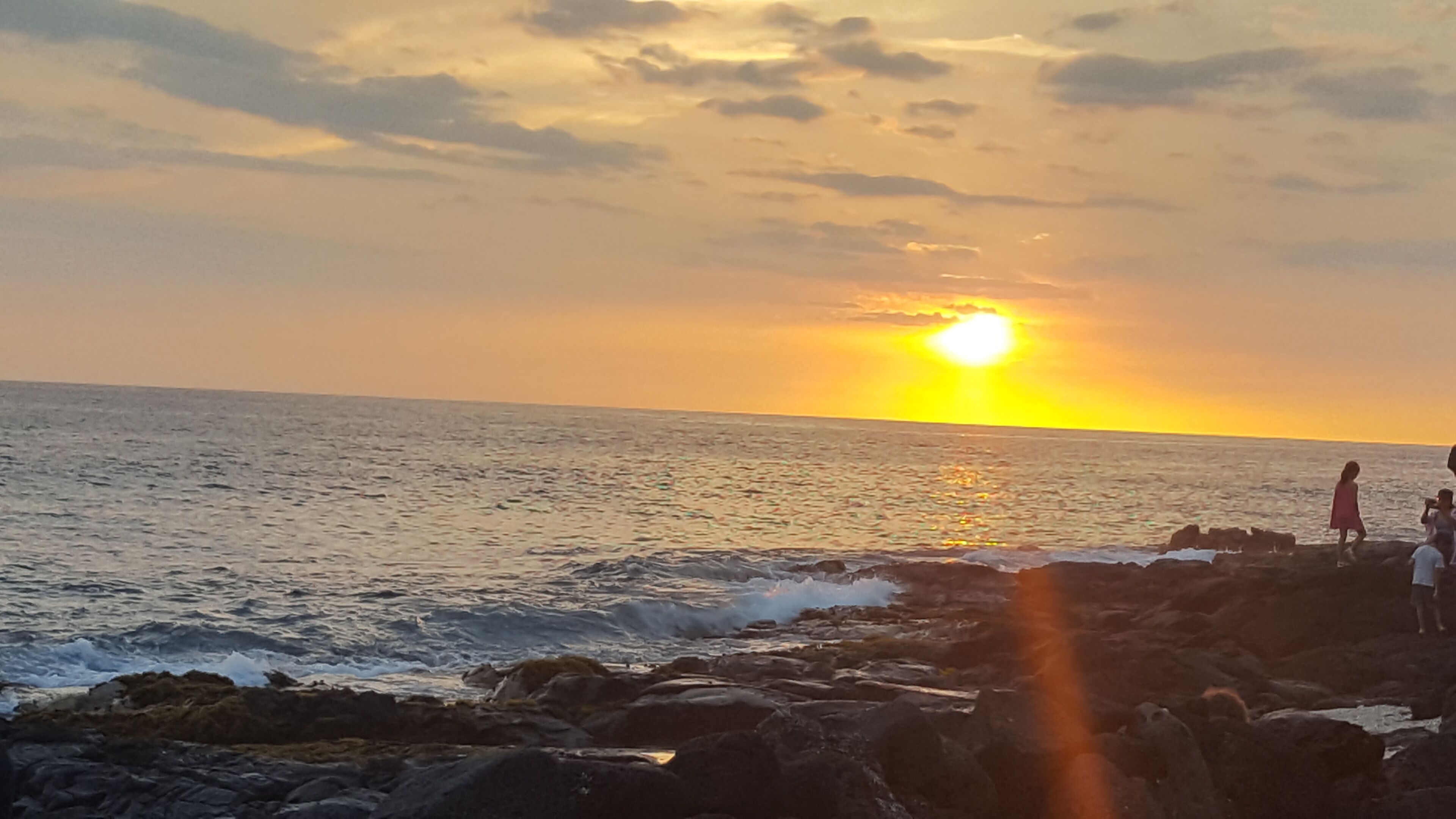 A sunset over the ocean with rocky terrain and a few people in the distance.