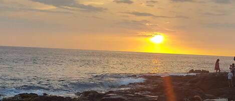 A sunset over the ocean with rocky terrain and a few people in the distance.