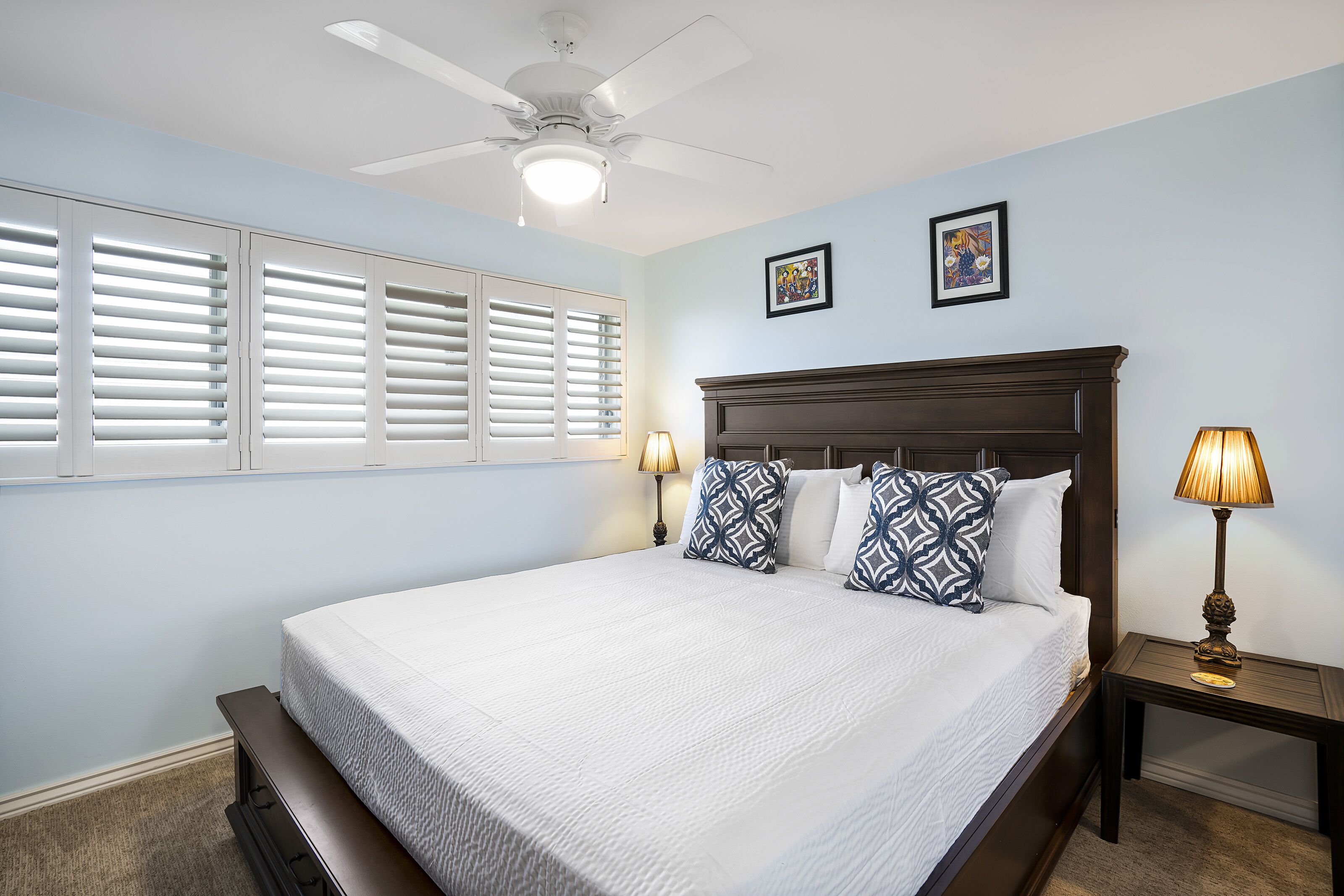 A bedroom with a large bed, two wall-mounted picture frames, a ceiling fan, and a nightstand with a lamp.