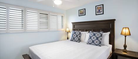 A bedroom with a large bed, two wall-mounted picture frames, a ceiling fan, and a nightstand with a lamp.
