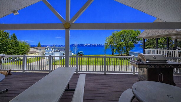 A covered outdoor area with a view of a lake and mountains, featuring picnic tables and a grill.