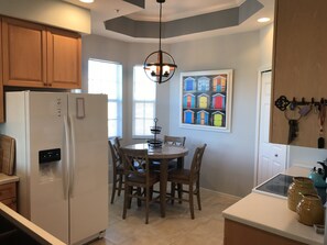 Una cocina con armarios de madera, un refrigerador blanco, una mesa de comedor con sillas, una lámpara colgante y una obra de arte de pared colorida.