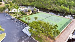 Un court de tennis entouré d’arbres et bordé par un plan d’eau, avec un parking et un bâtiment situés à proximité.