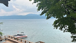 A wooden deck with kayaks, a paddleboard, and a yellow slide, overlooking a calm lake with boats and distant mountains.