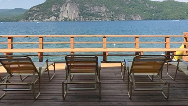 A wooden deck with metal chairs overlooking a body of water and a green hill in the distance.