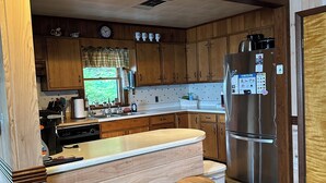 A modern kitchen with wooden cabinets, a stainless steel refrigerator, a bar area with stools, and a window with a view.