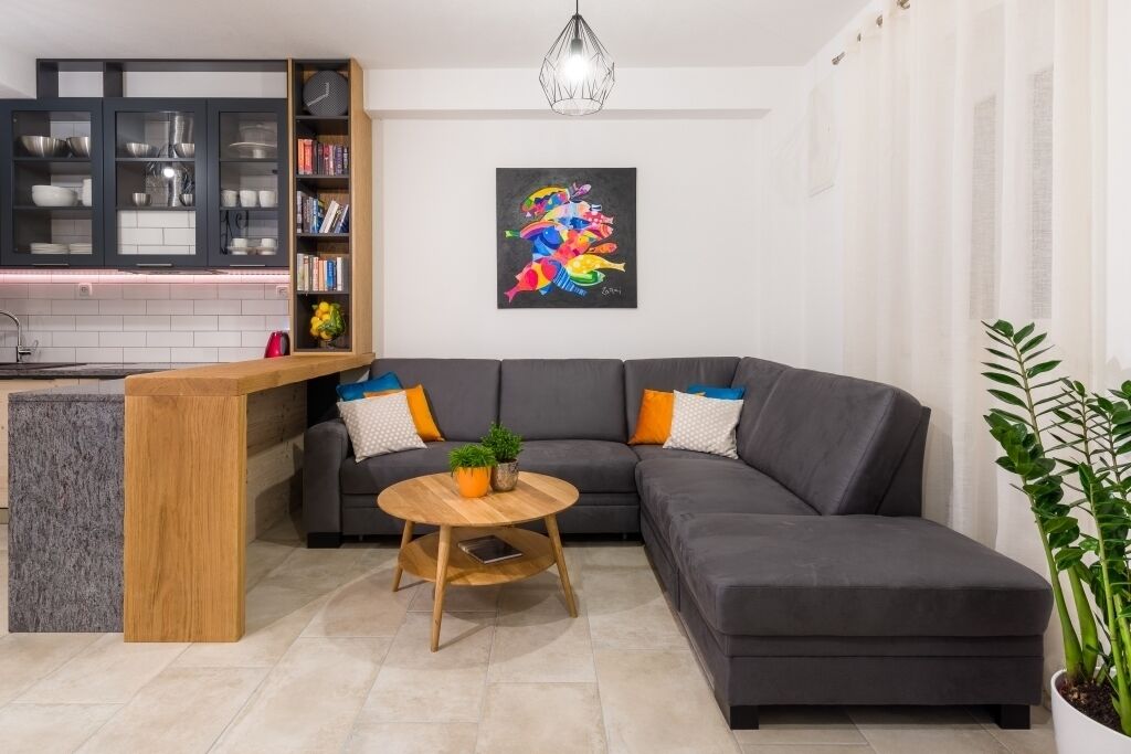 Uma sala de estar moderna com um sofá seccional cinza, uma mesa de centro de madeira e uma área de cozinha com armários escuros e bancada branca.