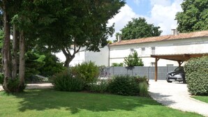 A white building with a tiled roof, a covered parking area, and a well-maintained lawn. - La Réserve, 2/4 person duplex gite, 9x4 pool, Saintes / Royan / Oléron axis (Luchat)