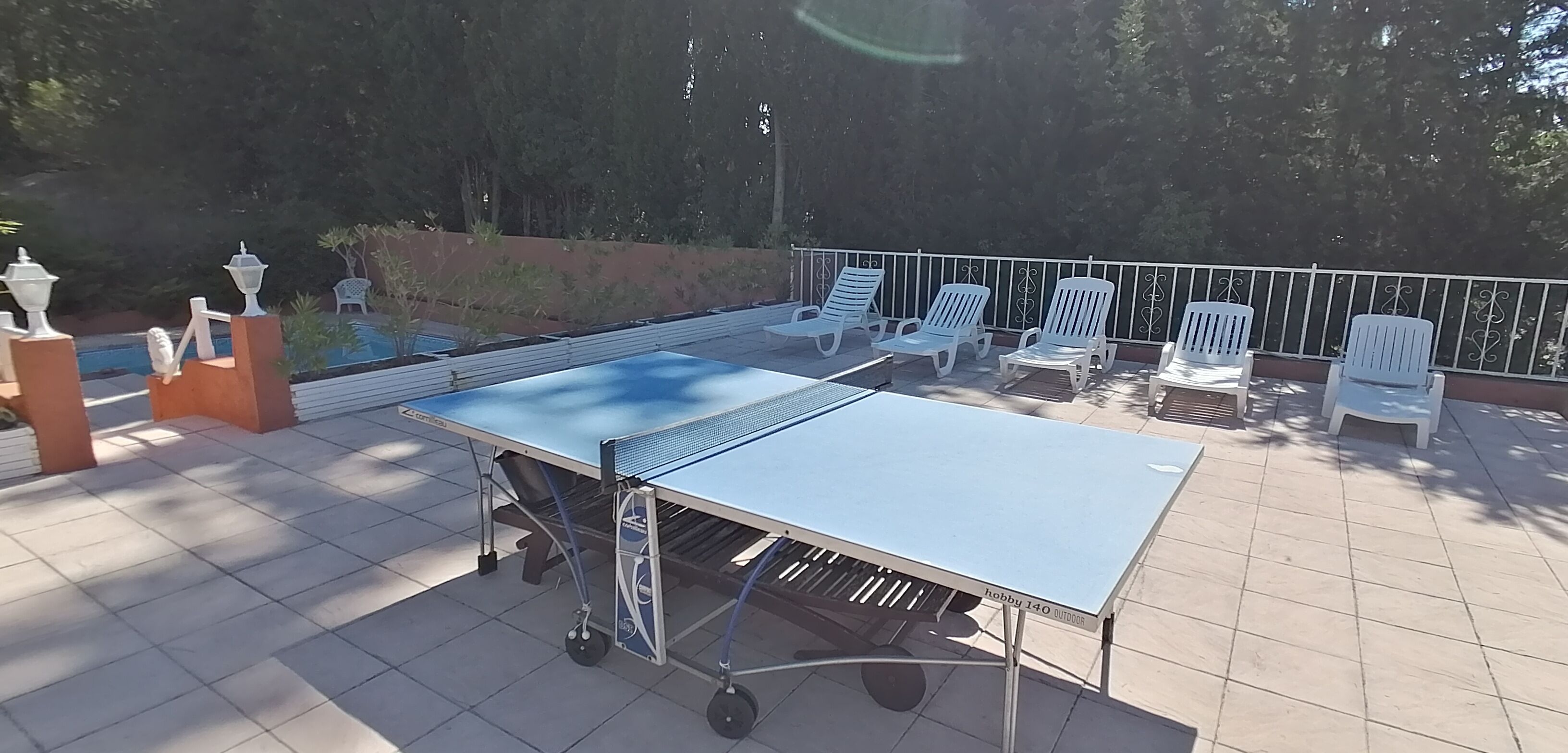 Une table de ping-pong d’extérieur, dotée d’un cadre en métal et de roulettes, entourée de chaises en plastique blanc.
