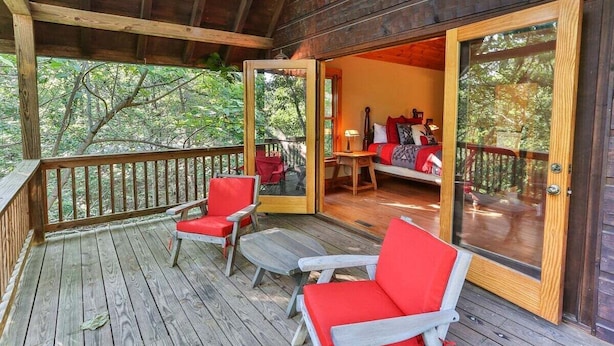A wooden deck with red chairs and a table, leading to a room with a bed and a window overlooking a forest.