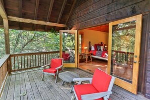 A wooden deck with red chairs and a table, leading to a room with a bed and a window overlooking a forest.