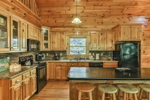 A kitchen with wooden cabinets, a central island with stools, a stove, microwave, refrigerator, and a window with a view of trees.