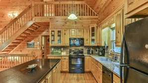 A wooden cabin interior with a kitchen, wooden staircase, and a large window.