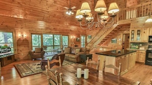 A spacious cabin with a wooden interior, a dining area with a large table, a kitchen with wooden cabinets, and a staircase leading to an upper level.