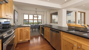 Une cuisine moderne avec des meubles en bois, un plan de travail noir, des appareils en acier inoxydable et de grandes fenêtres offrant une vue sur l’extérieur.