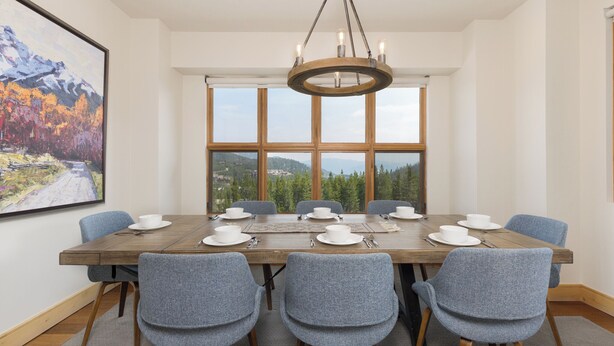 Une salle à manger avec une table en bois dressée pour six personnes, six chaises grises à revêtement rembourré, un lustre et une grande fenêtre donnant sur la montagne.