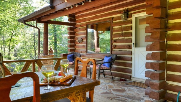 Eine Holzhütte mit einer Steinterrasse, Korbstühlen, einem Tisch mit einem Weinglas und Blick auf den Wald.
