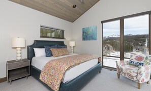 A bedroom with a large bed, two bedside lamps, a window with a view of mountains, and a chair with a ski-themed design. - Big Sky Vacation Rentals: Homestead 14 Claim Jumper (Big Sky)