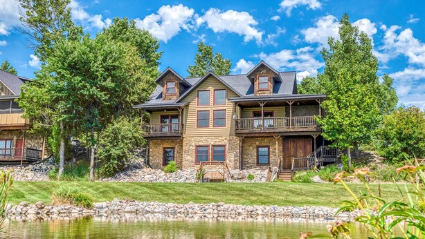 A large, multi-story house with a stone facade, multiple balconies, and a pond in the foreground.